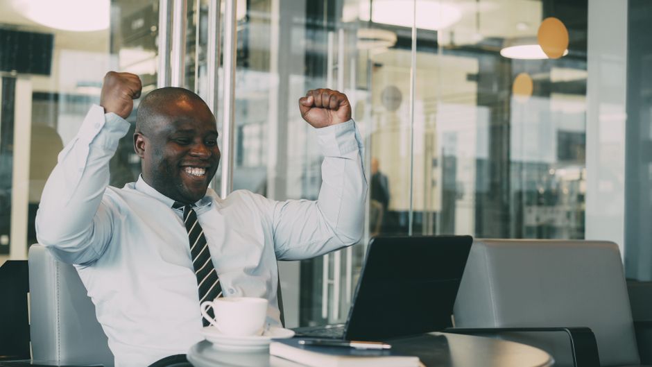 Happy businessman celebrating a success while working on a laptop in a modern office setting.